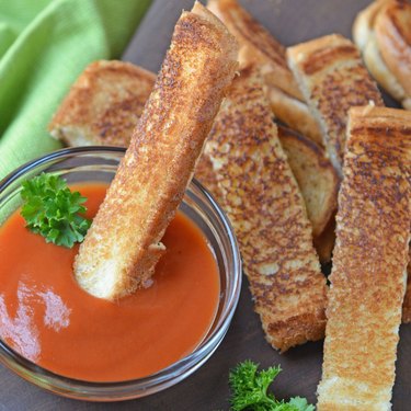 Grilled cheese sticks on a dark gray surface with one stick being dipped in a small glass bowl of tomato soup.
