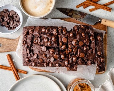 A loaf of chocolate pumpkin bread with chocolate chips on a wooden cutting board with a knife, cinnamon sticks, and a bowl of chocolate chips.