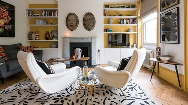 A living room with yellow and white walls and taupe window treatments, white chairs and a black and white rug.