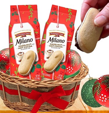 Two red bags of Milano Hazelnut Hot Cocoa in a basket with a red bow next to a hand holding a Milano cookie.