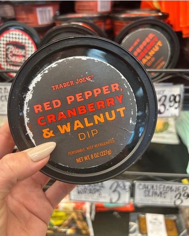 A white woman's hand holds a tub of Trader Joe's red pepper, cranberry and walnut dip in front of the dip aisle. The lid of the tub is black, and the text is coral, red and yellow