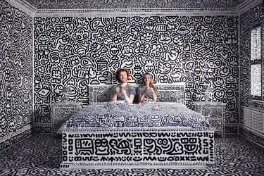 Sam Cox and his wife sitting on a doodled white bed in a completely doodled bedroom.