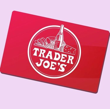 A red Trader Joe's gift card in front of a pink background. The Trader Joe's logo is white, with an image of bread, wine and cheese above it.