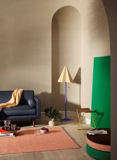A beige floor lamp with a blue stem in a beige living room next to a dark gray couch and a peach-colored carpet.
