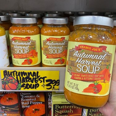 Hand holding a jar of Trader Joe's Autumnal Harvest Soup with more jars in the background on the store shelf.
