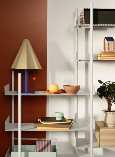 A beige table lamp with a blue stem on a white shelf that is stacked with other home goods like bowls, books, and plants.