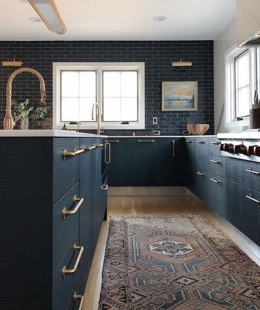 Dark teal kitchen cabinets, matching teal subway tile backsplash, oriental rug, brass fixtures, sink.