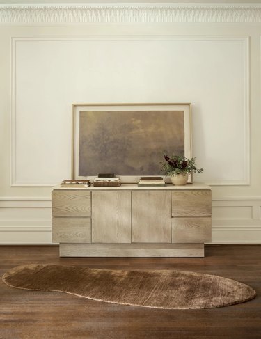 An irregular-shaped brown rug in front of a light gray wood dresser on dark hardwood floors.