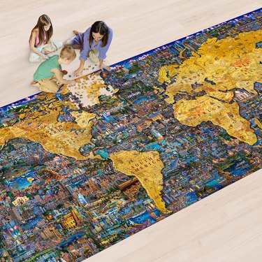 A white woman with dark brown hair sits on the floor with two children, a boy and a girl. They are putting together the world's largest puzzle, which lays on the floor in front of them. The puzzle is a map of the world, with picturesque scenes from around the world in place of where the ocean house be.