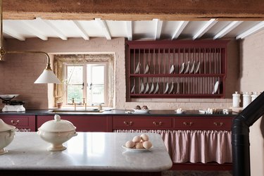 A kitchen with light pink brick walls and cranberry-colored cabinetry, along with a wall-mounted plate rack.