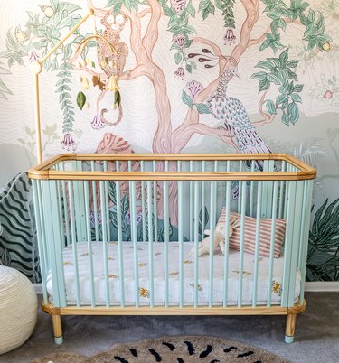 A crib with light blue bars and light wood on the bottom and top pieces, in front of a jungle wall mural in a nursery room.