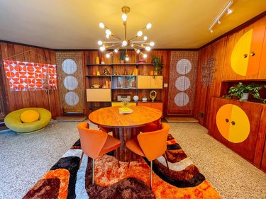 A '70s-inspired dining room with wood walls, white and brown flecked carpet, a starburst chandelier, a round wood table, and orange dining chairs.
