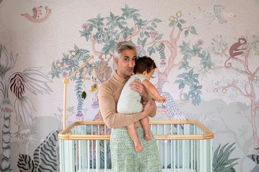 Tan France, a person with short gray hair, holding a baby with short brown hair, in front of a crib and jungle wall mural.