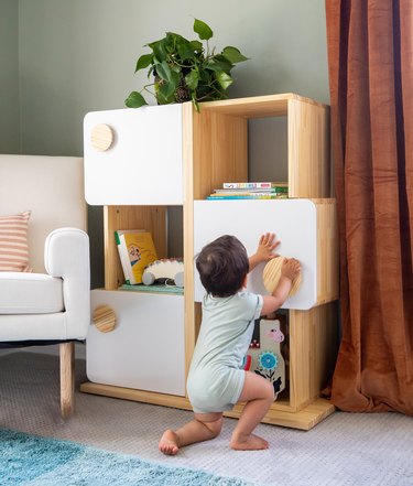A baby with short brown hair opening a cabinet in a light wood shelf that has three white cabinets and three open spaces.