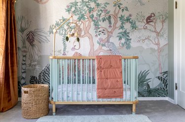 A jungle wall mural featuring trees, monkeys, birds, and a tiger behind a wood and light blue crib.