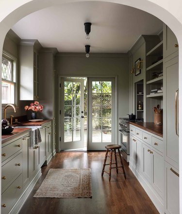 Light green kitchen with hardwood floors, built-in shelves, stool, wood counters, french doors.