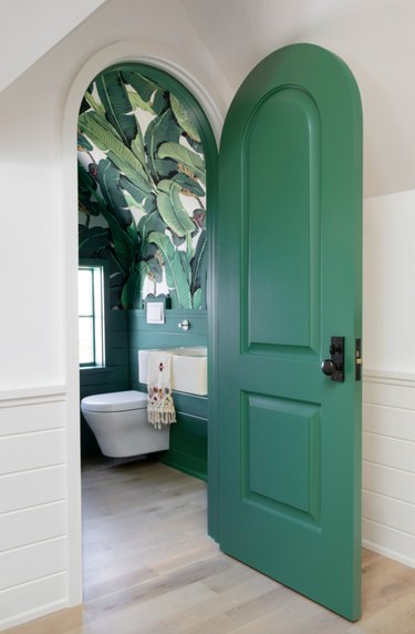 Green door with arch shape, bathroom with green palm wallpaper, wall mounted toilet, shiplap walls, sink, wood floors.