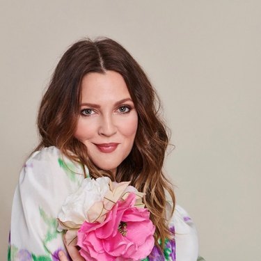 Drew Barrymore in a white top holding fake flowers in front of a beige wall.