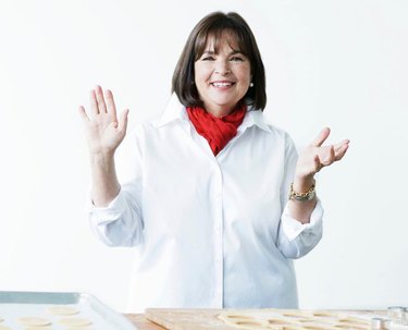 Ina Garten in a white shirt and red scarf in front of a kitchen counter with a white background.