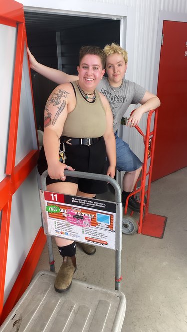 Sam Golub, a white person with short brown hair, and Cam Schroeder, a white person with short blonde hair, of Butch-4-Hire, holding a step ladder and hand truck.