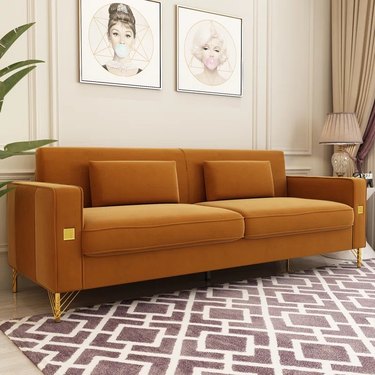 Wayfair's Velvet Square Arm Sofa in orange on a patterned carpet in front of a beige wall with drawings of Audrey Hepburn and Marilyn Monroe hanging on the wall.