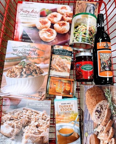 A Trader Joe's cart filled with Thanksgiving food, including Pastry bites, fried onions, cranberry sauce, turkey gravy, canned pumpkin, cornbread stuffing mix, a bottle of witch's brew, turkey stock, rustic apple pie and a tofu turkey