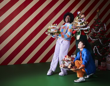 Three people holding piled plates of food on a green floor in front of a candy cane striped wall.