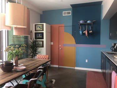 Modern kitchen with bold paint pattern on wall, concrete floors, dining table and chairs, pendant lamps, pots, rack, black cabinets.