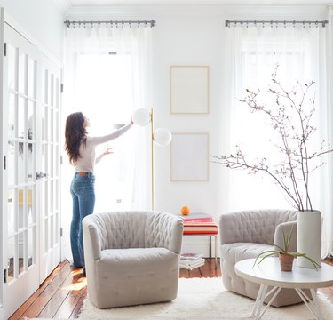 Woman adjusting curtains in white, bright room w/ modern furniture