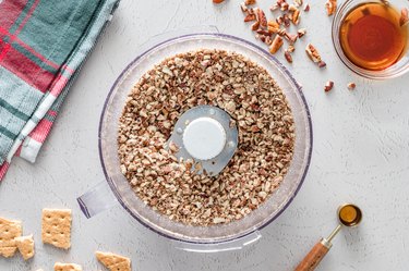 Chopped pecans in a food processor on a white counter next to a red and green plaid kitchen tea towel.