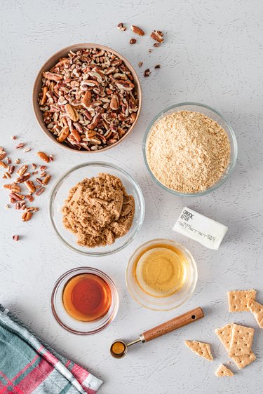 The pecans, graham cracker crumbs, dark brown sugar, vanilla extract, maple syrup, bourbon, and butter in glass bowls on a white counter.