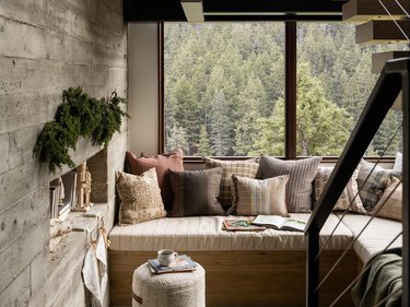 Sitting area with a built-in bench and muted warm toned pillows against a backdrop of large windows with a view of pine forest