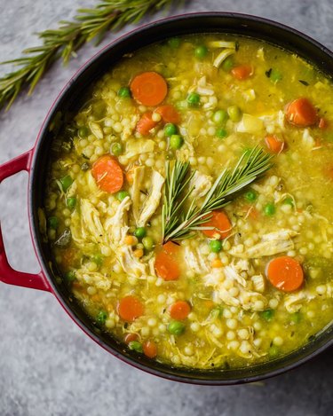 A red Dutch oven filled with chicken soap that contains carrots, peas, couscous, and rosemary.