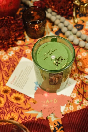 A green "night gardenia" scented candle on a table with an orange floral tablecloth.