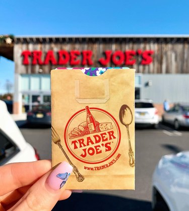 A person holding up a Trader Joe's gift card in front of a Trader Joe's store.