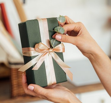 A person placing a sprig of eucalyptus in a light gold ribbon on a green present.