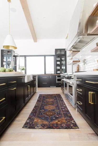Kitchen with black cabinets, light wood floors, oriental rug, brass fixtures, white subway tile.