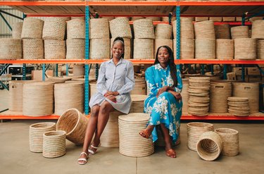 Mo and Michelle Mokone sitting on woven baskets in front of shelves of more woven baskets.