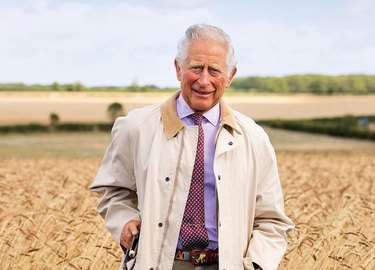 King Charles standing in a field in a tan trench coat.