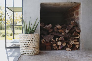 A woven basket with a plant next to a display of firewood in front of a white wall.