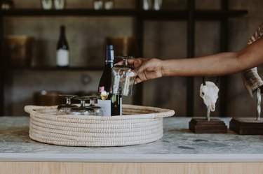 A hand placing a soap container on a woven tray next to a wine bottle and other dispensers.