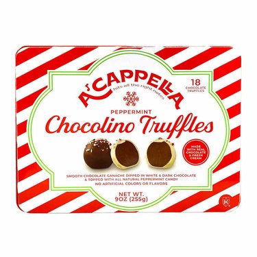 A red and white striped tin of Acappella Chocolino Truffles. Pictured is one chocolate truffle, with a white chocolate truffle sliced in half, with a dark chocolate inside.