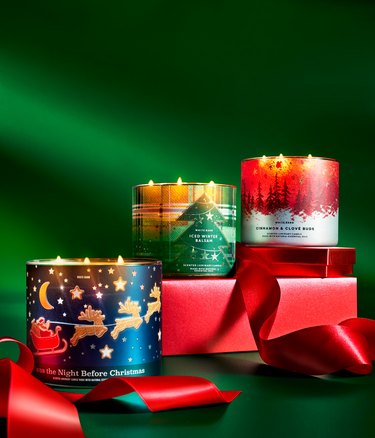 A stack of red presents against a green background with three Bath and Body Works 3-wick candles displayed: Twas the Night Before Christmas, Iced Winter Balsam, and Cinnamon and Clove Buds