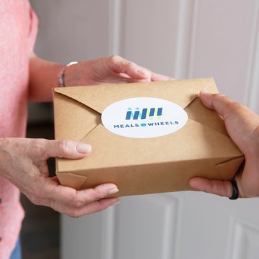 Hands giving another hand a to-go food box that reads "Meals on Wheels"