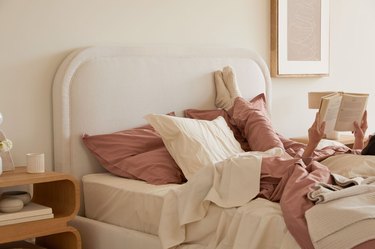 Bedroom with beige bedding and dark pink pillows with a person laying with their feet up reading a book.