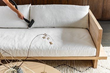 A person vacuuming the cushions of a white sofa.