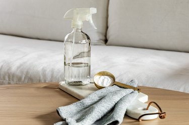 A spray bottle of vinegar, a tablespoon of baking soda, and a cloth sitting on a wood coffee table in front of a fabric couch.
