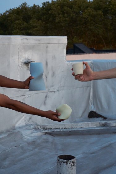 Three hands holding curvy vases and cups in front of a low cement wall outside.