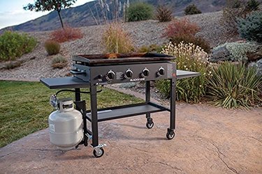 Blackstone 1554 4-Burner Flat-Top Gas Grill With Propane Canister on Patio in Landscaped Yard