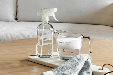 A measuring cup and spray bottle filled with white vinegar on a wood coffee table in front of a white fabric couch.